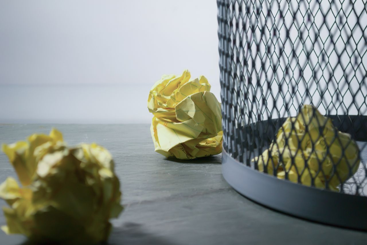 Close-up of crumpled yellow paper around a mesh trash bin, symbolizing rejected ideas or recycling.